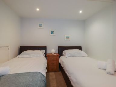 A bedroom with two single beds separated by a wooden nightstand with a lamp and three small pictures on the wall at St Saviours Padstow