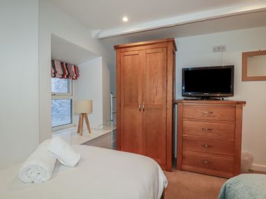 A bedroom with a bed, wooden wardrobe, chest of drawers with a TV on top, and a window with a striped blind at St Saviours in Padstow