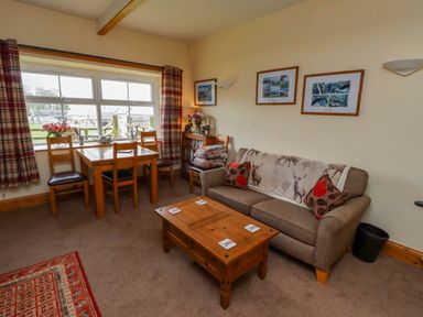 A living room with a wooden dining table and chairs next to a window with plaid curtains a sofa with cushions and a throw blanket a wooden coffee table and framed pictures on the wall at Folly View in Eglingham