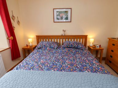 A bedroom with a wooden bed with floral bedding two bedside tables with lamps a wooden chest of drawers and red curtains at Folly View in Eglingham