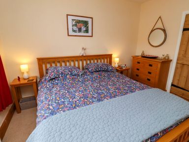 A bedroom with a wooden double bed with floral bedding two bedside tables with lamps a wooden dresser with a round mirror and wall art above the bed at Folly View in Eglingham