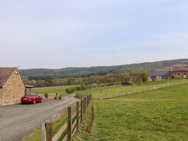 A gravel driveway with a red car next to a stone building and fenced green fields with distant trees and hills at Folly View in Eglingham