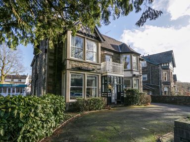 A stone building with large windows and a small balcony with bushes and trees in front at Fir Trees in Windermere