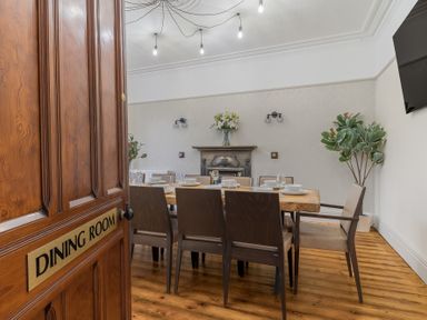 A dining room with a wooden table set with dishes and chairs near a fireplace and a potted plant at Fir Trees in Windermere