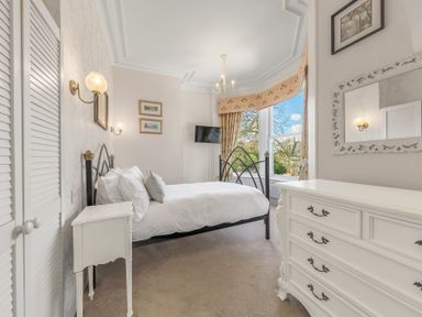A bedroom with a metal bed frame white bedding a white dresser a window with curtains and a wall-mounted television at Fir Trees in Windermere