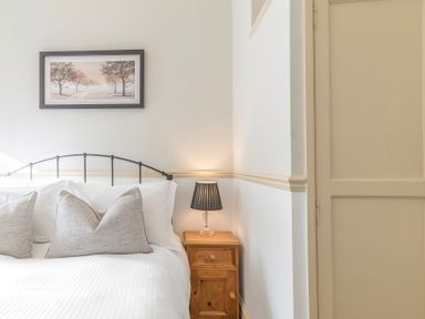 A bedroom corner with a bed white pillows a wooden bedside table with a lamp and a framed picture on the wall at Fir Trees in Windermere