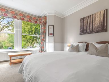 A bedroom with a double bed pillows a wooden bench and large windows with floral curtains at Fir Trees in Windermere