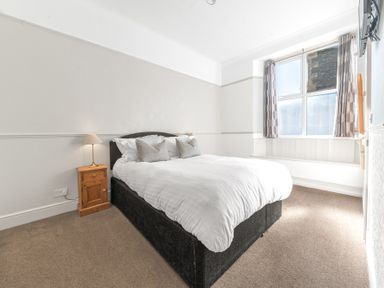 A bedroom with a double bed two pillows a wooden bedside table with a lamp a window with curtains and a wall-mounted television at Fir Trees in Windermere