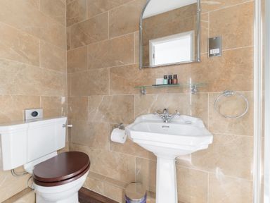 A bathroom with a white toilet with a dark wooden seat a pedestal sink a mirror and toiletries on a glass shelf at Fir Trees in Windermere