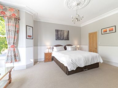 A bedroom with a double bed two bedside tables with lamps paintings on walls and a window with floral curtains at Fir Trees in Windermere