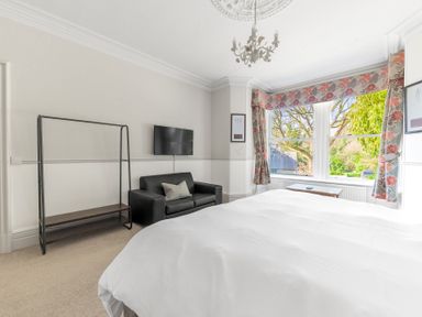 A bedroom with a bed a black loveseat a wall-mounted TV a clothes rack and floral curtains at Fir Trees in Windermere