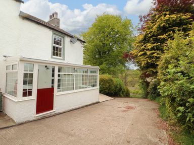 Exterior view of a white house with a red door and glass porch next to a driveway and trees at Kirkland Howe Cottage in Arlecdon