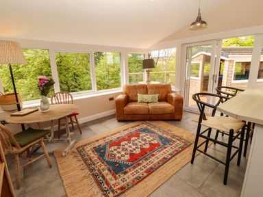 A sunroom with a leather sofa a round wooden table with chairs and bar stools at Kirkland Howe Cottage in Arlecdon