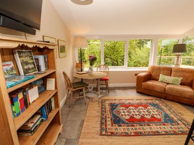 A living room with a wooden bookshelf a round table with two chairs a brown leather sofa and large windows overlooking trees at Kirkland Howe Cottage in Arlecdon