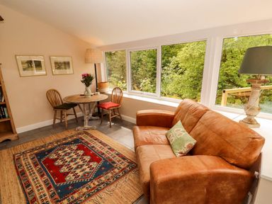 A living room with a leather sofa a small round wooden table with chairs a floor lamp and large windows showing greenery outside at Kirkland Howe Cottage in Arlecdon