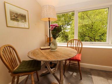 A wooden round table with a flower vase and lamp next to two wooden chairs with seat cushions by a window with a view of trees at Kirkland Howe Cottage in Arlecdon