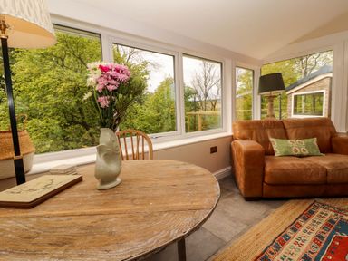 A room with a round wooden table with a vase of flowers a brown leather sofa and large windows at Kirkland Howe Cottage in Arlecdon