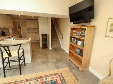 A kitchen area with a sink and chair and a wooden bookshelf with books beneath a wall mounted tv at Kirkland Howe Cottage in Arlecdon