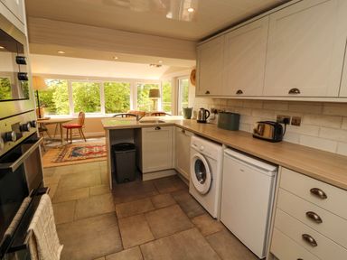A kitchen with white cabinets and appliances next to a dining area with chairs and windows showing trees at Kirkland Howe Cottage in Arlecdon