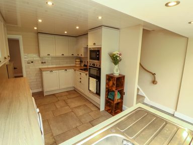 A kitchen with white cabinets a built in oven a wooden shelf with flowers and towels and a sink in the foreground at Kirkland Howe Cottage in Arlecdon