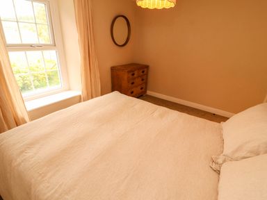 A bedroom with a bed covered in cream bedding a wooden chest of drawers a round mirror and window with curtains at Kirkland Howe Cottage in Arlecdon