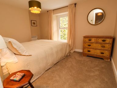 A bedroom with a bed a wooden chest of drawers a round mirror a side table and a window with curtains at Kirkland Howe Cottage in Arlecdon