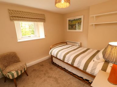 A bedroom with a single bed striped bedding a chair with a fur cover a window and wall art at Kirkland Howe Cottage in Arlecdon