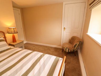 A bedroom with a striped bedspread a bedside table with a lamp and a striped chair with a fur cover at Kirkland Howe Cottage in Arlecdon