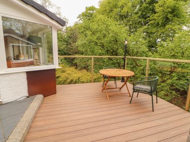 An outdoor deck with a wooden round table and two green chairs surrounded by glass railings and trees at Kirkland Howe Cottage in Arlecdon