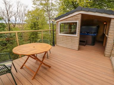 A wooden deck with a round wooden table two chairs a wooden shed with a hot tub and trees in the background at Kirkland Howe Cottage in Arlecdon