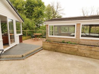 An outdoor patio area with a small wooden table and two chairs next to a building with large windows at Kirkland Howe Cottage in Arlecdon