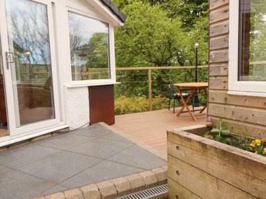 A wooden deck with a round table and chairs next to a house with large windows and a planter box with plants at Kirkland Howe Cottage in Arlecdon