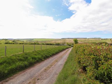 A dirt road running through green fields with wooden fence posts and hedges under a partly cloudy sky at Kirkland Howe Cottage in Arlecdon