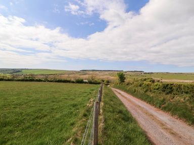 A rural dirt road next to a wire fence with green fields and hills under a partly cloudy sky at Kirkland Howe Cottage in Arlecdon