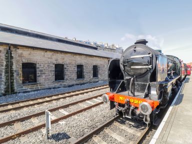 A steam locomotive at a train station platform with a stone building alongside Duchess of Hamilton @ Engine Shed in Whitby