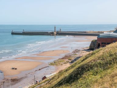 A beach with a pier extending into the sea and a grassy hill with a building at Duchess of Hamilton @ Engine Shed in Whitby