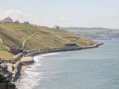 A coastal scene with colorful beach huts along a curved promenade and houses on a hillside at Duchess of Hamilton @ Engine Shed in Whitby