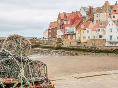 A stack of lobster pots on a stone pier with a sandy beach and houses with red roofs in the background at Duchess of Hamilton @ Engine Shed in Whitby