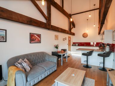 A living area with a gray sofa, a wooden table, two chairs, a kitchen island with two bar stools, and exposed wooden beams on the ceiling at Duchess of Hamilton @ Engine Shed in Whitby