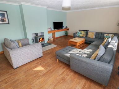 A living room with a gray sectional sofa a wooden coffee table a wood-burning stove and a wall-mounted television at Carreg Yr Onnen in Penparc