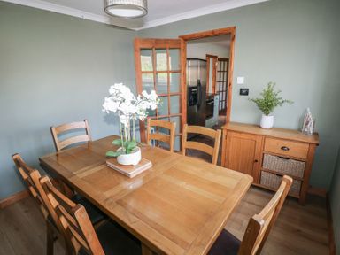 A dining room with a wooden table and six chairs a cabinet with plants and decor and a door leading to a kitchen at Carreg Yr Onnen in Penparc