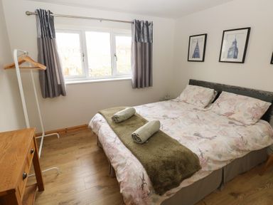 A bedroom with a double bed with floral bedding two rolled towels a wooden side table a clothes rack and three framed pictures on the wall at Carreg Yr Onnen in Penparc