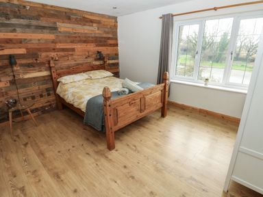 A bedroom with a wooden double bed wood panel wall beside a window with curtains and a small round glass side table at Carreg Yr Onnen in Penparc