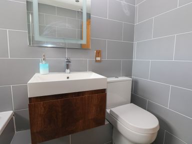 A bathroom with a white sink and wooden cabinet next to a white toilet and a rectangular mirror on the tiled gray wall at Carreg Yr Onnen in Penparc