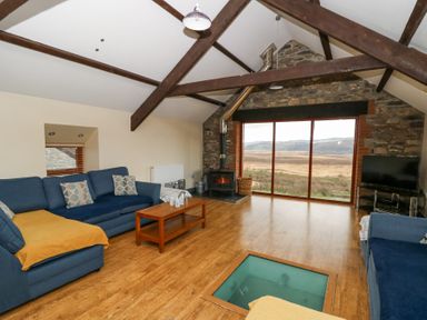 A living room with blue sofas a wood stove large window with a view and wooden ceiling beams at Llwyngwinau Isaf Barn Tynreithyn near Tregaron