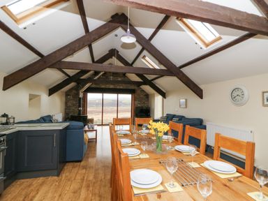 A dining area with a wooden table set with plates and glasses next to a living room with blue sofas a stove and large windows at Llwyngwinau Isaf Barn in Tynreithyn near Tregaron