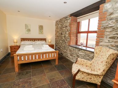 A bedroom with a wooden bed two bedside tables a floral armchair and a window with stone and brick walls at Llwyngwinau Isaf Barn in Tynreithyn near Tregaron