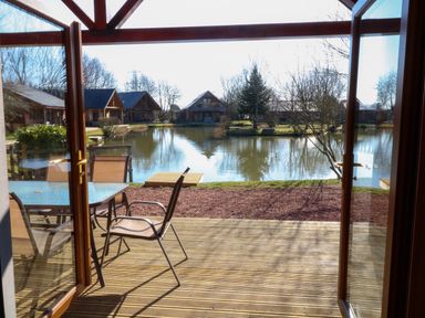 A patio with table and chairs overlooking a lake with cabins and trees at The Green Monkey in North Hykeham
