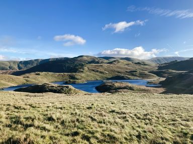 A grassy field with a lake and hills under a blue sky at Low Wood View in Hartsop