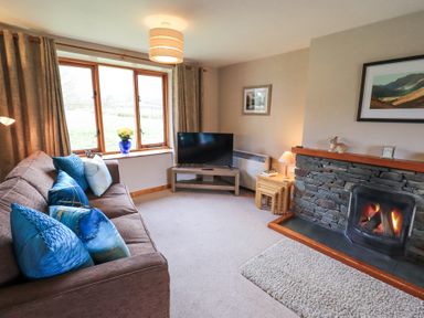 A living room with a brown sofa with blue cushions a stone fireplace a television on a stand and a window with curtains at Low Wood View in Hartsop
